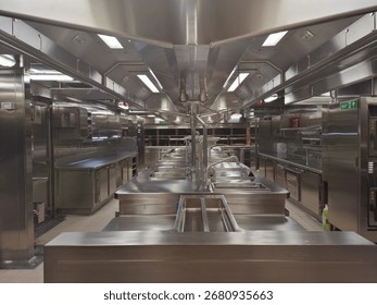 A clean and empty professional kitchen with a sleek, symmetrical design. The space is dominated by reflective stainless steel counters, sinks, and industrial-grade equipment under bright 