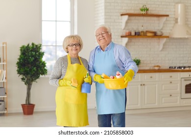 A Clean Home Is So Much Better. Happy Smiling Senior Couple In Yellow Aprons And Gloves Holding Cleaning Tools And Looking At Camera After Tidying Up Their House Or Studio Apartment Together