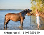 The clean horsewoman and her dirty bay mare are on a riverbank in summer.