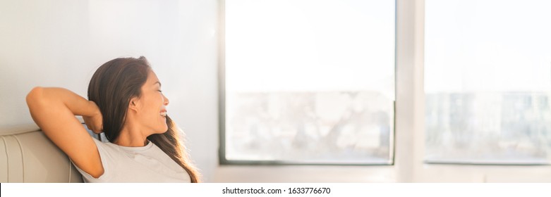 Clean indoor air happy Asian woman breathing at home from air purifier sitting in sofa looking out the window panoramic banner.
