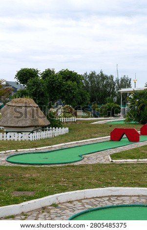 A clean and inviting outdoor mini golf course featuring multiple putting greens, white concrete borders, stone pathways, and playful obstacles including a small red bridge. The area is surrounded by lush greenery, trees, shrubs, and decorative elements such as a thatched-roof hut and picket fence, creating a friendly and family‑oriented atmosphere. Ideal for commercial use in advertising, leisure industry promotions, tourism materials, recreation concepts, family entertainment, sports activities, and outdoor lifestyle projects. 