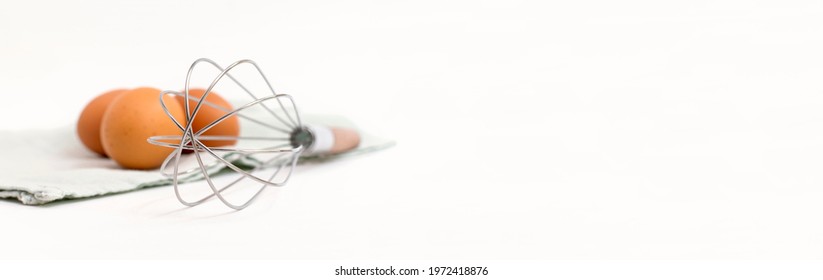 Clean metal whisk and raw brown eggs on a kitchen towel are located on the right, light banner background, copy space for text. Culinary mixer, whipping egg white and yolk, preparing and mixing cream.
