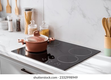 Clean new and black induction stove with control panel near marble countertop on white kitchen. Metal saucepan and jars with products on cooking surface. Cookery and homemade food concept