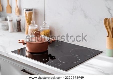 Clean new and black induction stove with control panel near marble countertop on white kitchen. Metal saucepan and jars with products on cooking surface. Cookery and homemade food concept