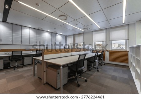 A clean office space shows empty desks arranged in rows. Bright lights are on and sunlight enters through the windows. The room is tidy and offers a workspace for employees.