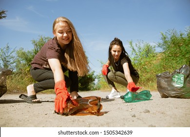 Clean Our Home Together. Group Of Volunteers Tidying Up Rubbish On Beach In Sunny Day. Young Women Take Care Of Nature And Environment, Taking Bottles And Packs Away. Concept Of Ecology.