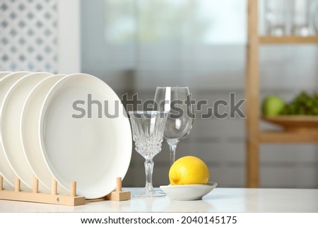 Clean plates in dish drying rack, glasses and lemon on white table indoors