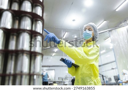 Clean room worker using tablet to inspect stacked cans in canned fish factory, highlighting food processing, packaging quality control, hygiene standards, industrial safety, production monitoring.