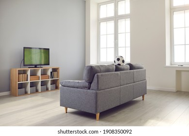 Clean, Tidy, Minimalist Living-room Interior With Sofa, Plastic Windows And TV With Soccer Match On. Empty Living Space Or Rec Room With Television Set, Books And Soft Grey Couch With A Football On It