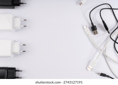 A clean, top-down studio shot features an organized array of essential electronic charging accessories on a bright white background. On the left, a neat column of black and white two-pin European power adapters provides a visual anchor. The right side displays a tangle of black and white USB cables, including USB-A, USB-C, and Micro-USB connectors, ready for various devices. This well-lit image emphasizes modern connectivity and power solutions for smartphones, tablets, and other gadgets, conveying order and technological readiness.