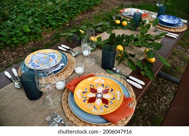 Clean, Unused Plates On A Picnic Table Outside. There Is A Lemon Garland. There Are Also Candles And Silverware. The Table Is Long.