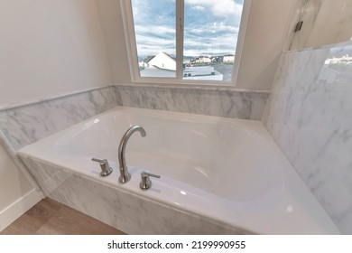 Clean White Built In Bathtub Of Home With View Of Neighborhood Houses. The Tub Is Inside A Residential Bathroom With Tiled Wall And Cloudy Sky View Through A Window.