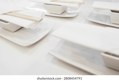 Clean white table setting at a restaurant, featuring ceramic plates, small bowls, folded napkins, and wooden chopsticks, ready for a meal