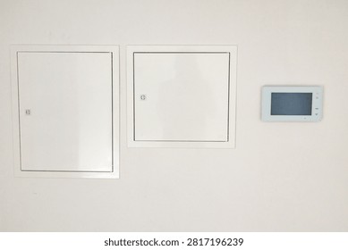 A clean white wall features two flush-mounted access doors and a small electronic interface device mounted nearby