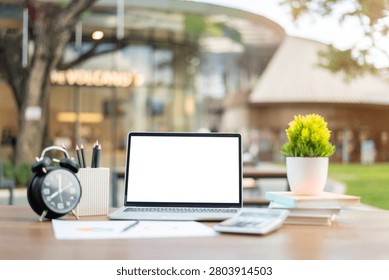 A clean workspace mockup featuring an open laptop with blank screen, alarm clock, pencils, calculator, books, and potted plant on a wooden desk outdoors.
