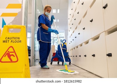 A cleaner with a mask on her face cleans the floor with the mop.Caution wet floor sign close up - Shutterstock ID 1812615982