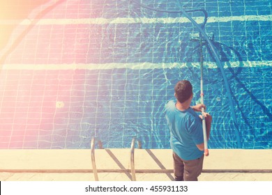 Cleaner of the swimming pool . Man in a blue t-shirt with a long hose near the pool, sunny
