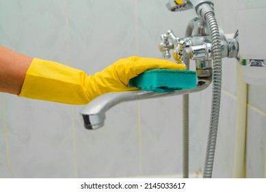 Cleaning bathroom, hand in yellow rubber glove washing chrome faucet with sponge. Close-up, selective focus.