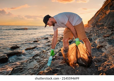 Cleaning Beach From The Pollution. Volunteer Picking Up A Plastic Bottle. In The Background, The Sea And The Sunset. Copy Space. The Concept Of Environmental Conservation.