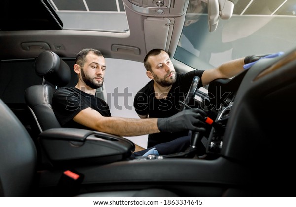 Cleaning of car interior at auto detailing service.
Two professional male workers, wearing black clothes and protective
gloves, cleaning control panel of the car with microfiber cloth and
soft brush