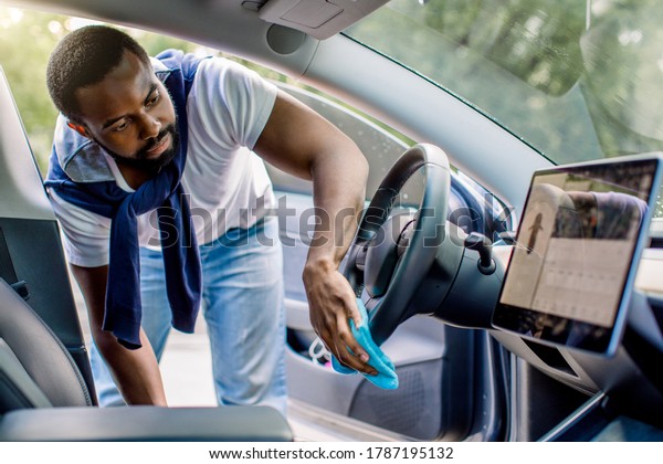 Cleaning of car\
interior, detailing concept. Young African man in casual wear\
washes a car interior, steering wheel with blue microfiber clothes,\
in a outdoor wash self\
service