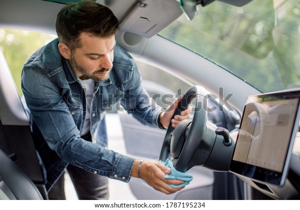 Cleaning of car\
interior, detailing concept. Young Caucasian man in casual wear\
washes a car interior, steering wheel with blue microfiber clothes,\
in a outdoor wash self\
service
