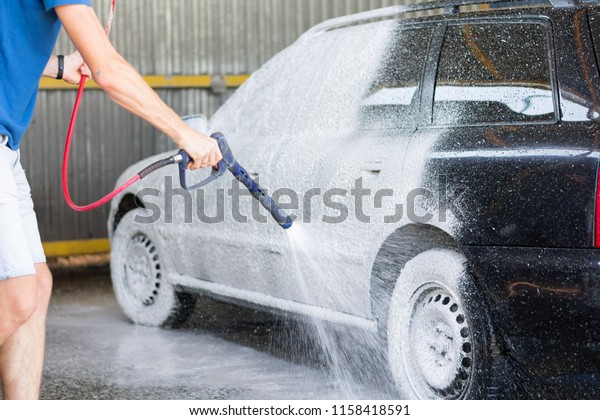 Cleaning Car Using High Pressure Water. Man washing\
his car under high pressure water in service.  Washing car with\
soap
