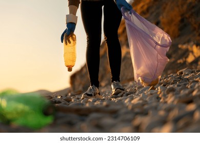 Cleaning of coastal areas from pollution. A volunteer walks along a pebble beach with a garbage bag and collects plastic bottles. Bottom view. The concept of preserving a sustainable ecosystem. - Powered by Shutterstock