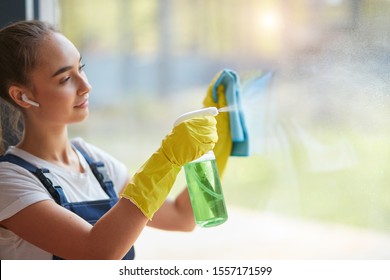 Cleaning Concept. Young Caucasian Girl With Blue Napkin Cleaning Panoramic Window At Home. Washing Windows With Spray