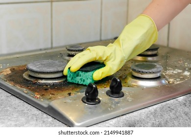 Cleaning Dirty Gas Stove From Grease, Food Leftovers Deposits. Woman's Hand In Protective Glove With Sponge Rag And Detergent Washing Kitchen Stove. Home Cleaning Service Concept.