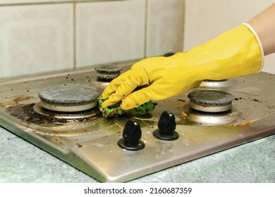 Cleaning Dirty Gas Stove From Grease, Food Leftovers Deposits. Woman's Hand In Protective Glove With Sponge Rag And Detergent Washing Kitchen Stove. Home Cleaning Service Concept.