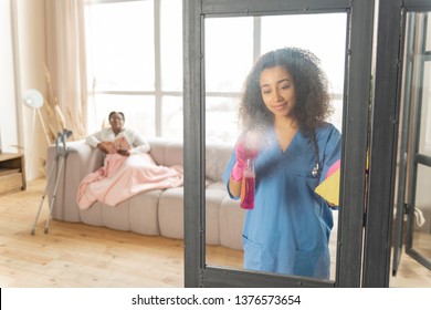 Cleaning Glass Door. Dark-skinned Private Nurse Cleaning The Glass Door In The Living Room Of Patient