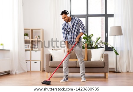 cleaning, housework and housekeeping concept - indian man in headphones with broom sweeping floor at home