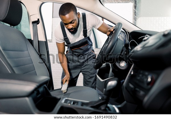 Cleaning of interior of the car with brush,\
car detailing service. Young pleasant black man doing car interior\
and seat cleaning with special\
brush