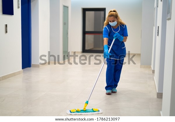 A cleaning lady with the mask on her face mops\
the floor in an office\
building