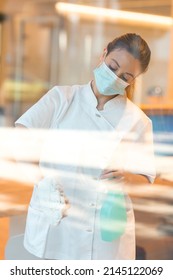 Cleaning Lady Using A Face Mask While Cleaning The Window Of A Spa Or Health Center.