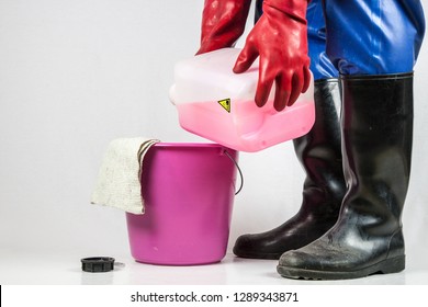 A cleaning person in a blue rubber protective suit, with  red rubber gloves and black rubber boots tilts a red, corrosive industrial cleaner in a plastic bucket.