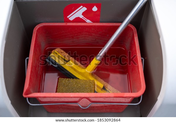 Cleaning set with squeegee, sponge and water for\
car windscreens in a red bucket at a gas station , selected focus,\
narrow depth of field