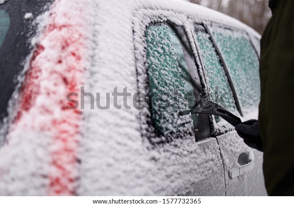 Cleaning the side car\
windows of snow with ice scraper before the trip. Man removes ice\
from car windows. Male hand cleans car with special tool at snowy\
frosty winter day.