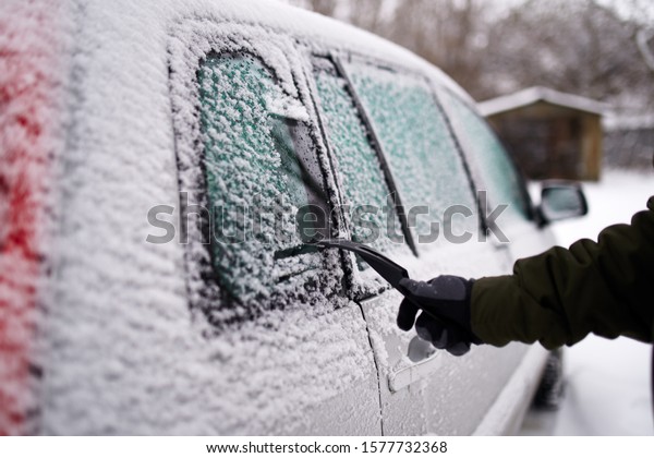 Cleaning the side car
windows of snow with ice scraper before the trip. Man removes ice
from car windows. Male hand cleans car with special tool at snowy
frosty winter day.
