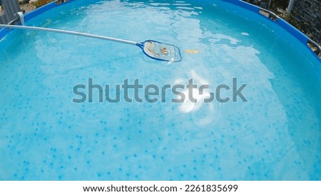 Cleaning the surface of the pool water from leaves with a net skimmer. View from above, wide angle. Beautiful flowers grow around the pool. Maintenance of a frame pool with a net skimmer