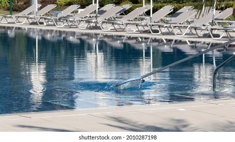 Cleaning swimming pool with skimmer net equipment in public water park of seaside resort summer morning, white sun loungers and umbrellas in background. Hospitality and tourism business concept.