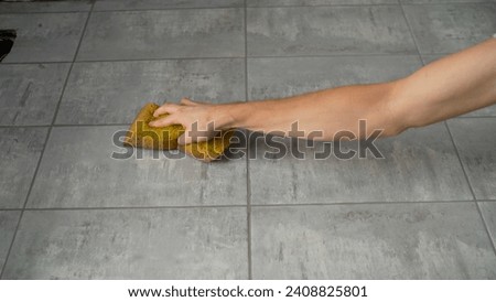 Cleaning tiles after renovation, hand with sponge close-up. A worker cleans tiles after laying them in the bathroom. A human hand uses a yellow sponge on a ceramic tile floor.