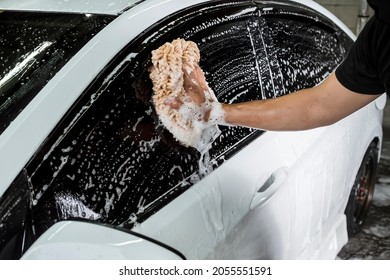 Cleaning the window of a white car with a microfiber wash mitt soaked in shampoo to lift grime and dirt. At a carwash or auto detailing shop. - Shutterstock ID 2055551591