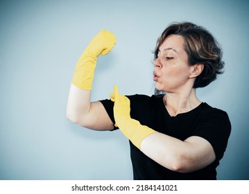 A Cleaning Woman In A Black T-shirt Looks At The Biceps On The Background. Vintage Woman Portrait. Woman In Yellow Rubber Gloves