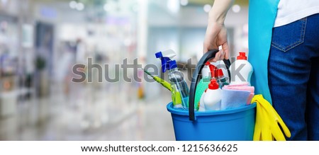 A cleaning woman is standing inside a building holding a blue bucket fulfilled with chemicals and facilities for tidying up in her hand.