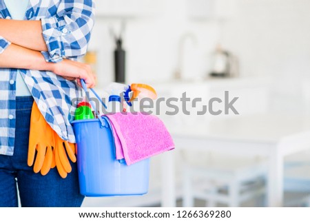 A cleaning woman is standing inside a building holding a blue bucket fulfilled with chemicals and facilities for tidying up in her hand