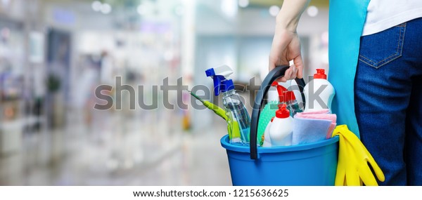 A cleaning woman is standing inside a building\
holding a blue bucket fulfilled with chemicals and facilities for\
tidying up in her hand.