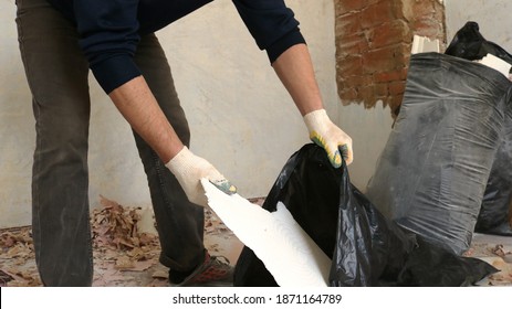 A Cleaning Worker In A Dark Uniform Collects Construction Waste With His Gloved Hands In A Cluttered Repair Room, Manual Collection Of Garbage And Trash In Large Black Bags