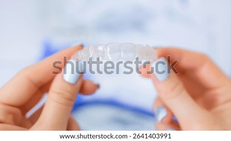 A clear aligner held by a pair of hands with light blue painted nails, orthodontic treatment to improve dental alignment and offer a transparent alternative to traditional braces.
